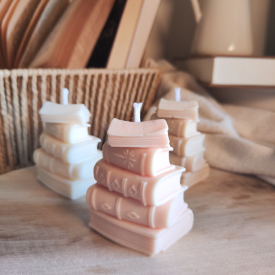 Velas decorativas em forma de pilhas de livros sobre superfície de madeira com cesta de vime e jarro branco ao fundo