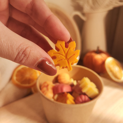 Mão segurando peça laranja em forma de folha com recipiente de papel cheio de decorações em forma de abóbora e folha no fundo