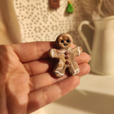 Boneco de gengibre decorativo marrom com detalhes brancos segurado por mão em fundo claro com padrão e enfeites natalícios.