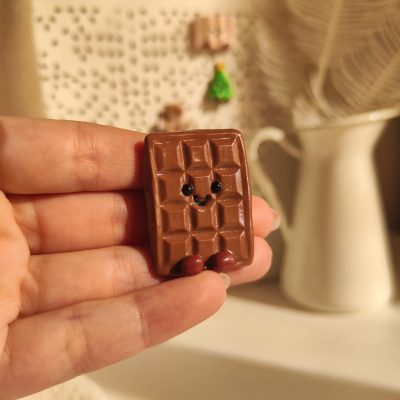Miniatura de barra de chocolate castanho com face e pés, segurada entre dois dedos.