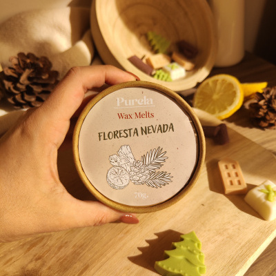 Embalaigem redonda de cera aromática Purela com texto FLORESTA NEVADA e peças em forma de árvores e objetos natalícios numa mesa de madeira