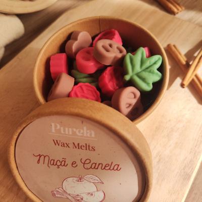 Recipiente com wax melts coloridos em forma de folhas e pretzels, etiqueta com texto Purela Wax Melts Maçã e Canela 70g sobre madeira