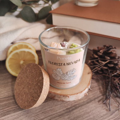 Vela aromática em vidro com tampa de cortiça e etiqueta FLORESTA NEVADA, decorada com limão, folhas verdes, livro e vela ao fundo