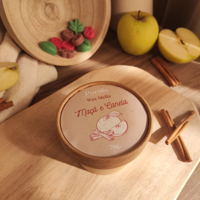 Caixa redonda de papel com label Purela Wax Melts Maçã e Canela 70g em mesa de madeira com paus de canela, maçãs e peça decorativa.