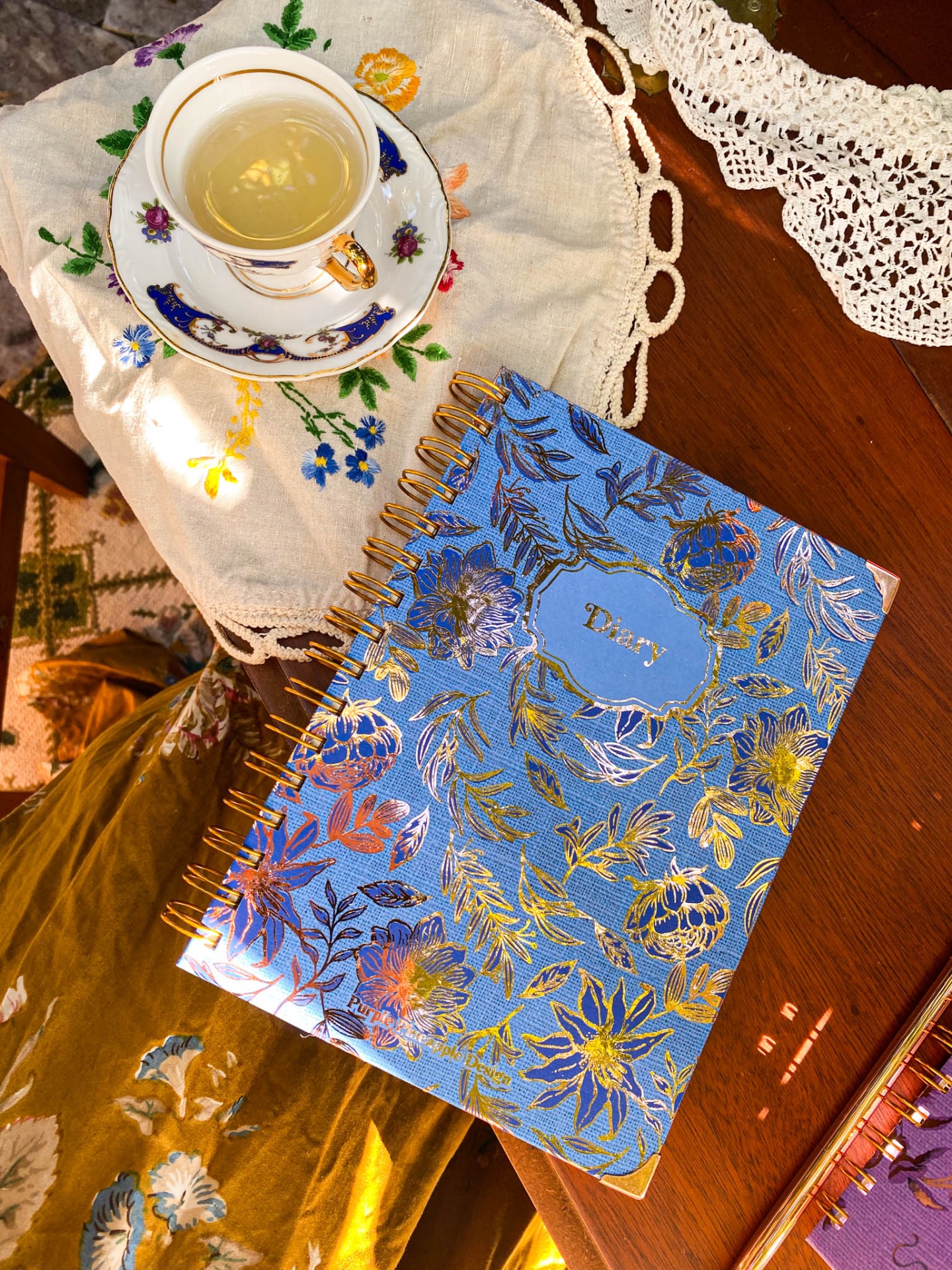 Caderno azul com decoração dourada e espiral dourada ao lado de chá num conjunto branco com azul e dourado