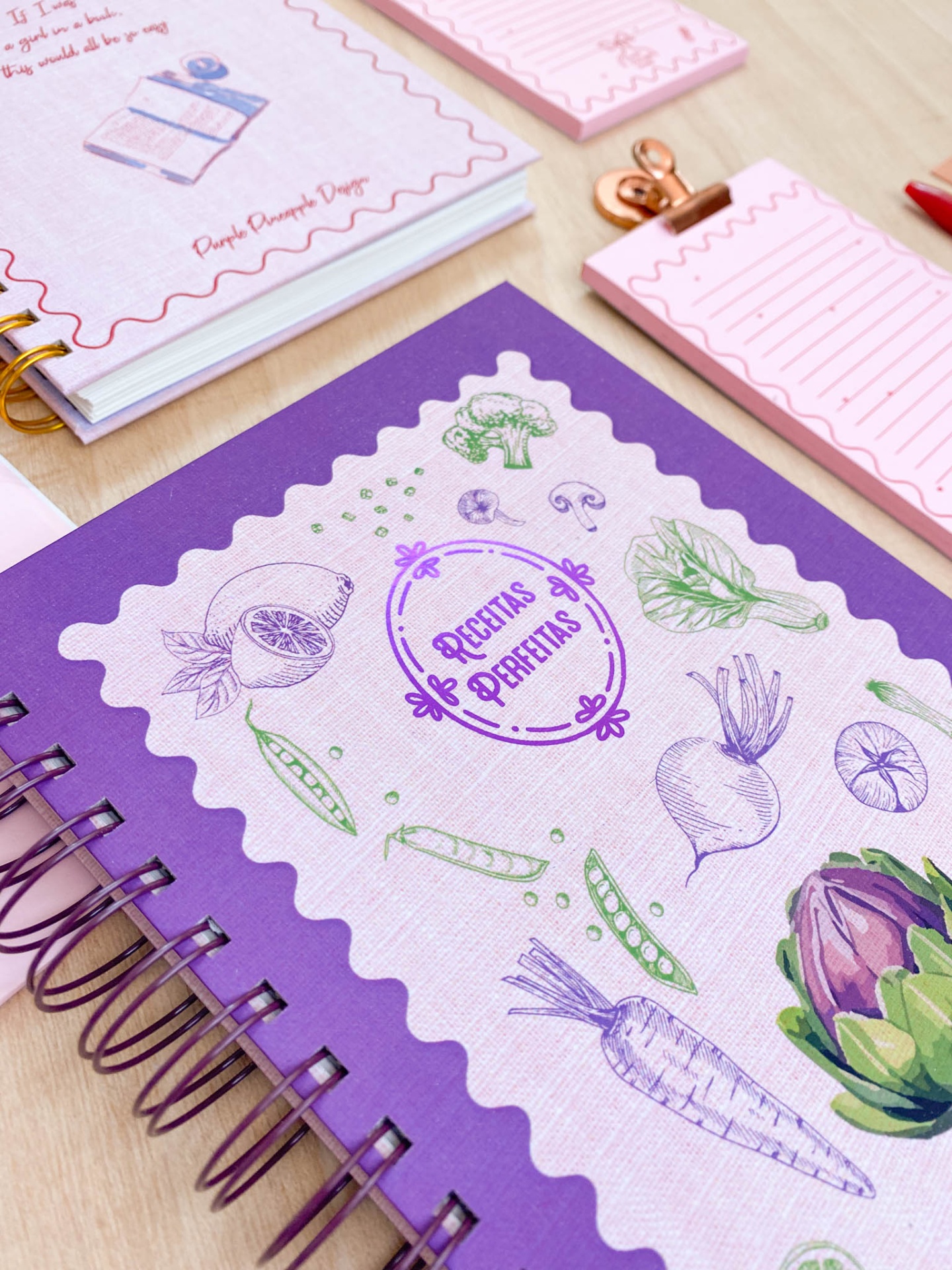 Caderno roxo de receitas com ilustrações de legumes e blocos de notas rosa com linhas e clips sobre mesa de madeira