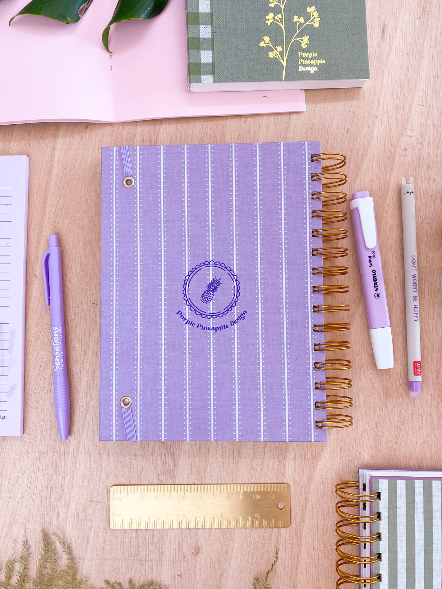 Caderno espiral lilás com padrão riscado branco e selo abacaxi, ao lado caneta roxa, marcador lilás e régua dourada sobre superfície de madeira.