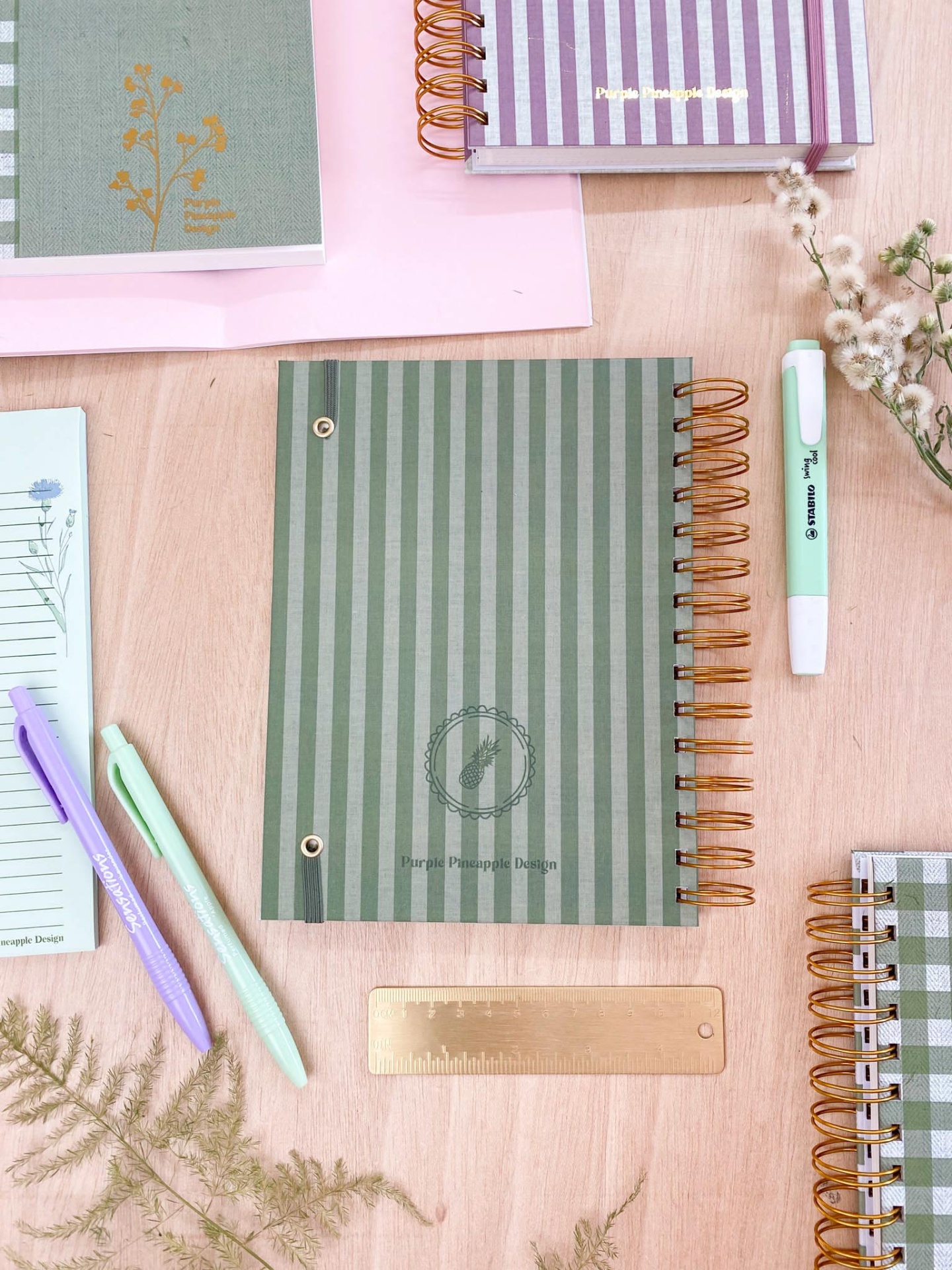 Cadernos espiralados com capas listradas, canetas coloridas e régua numa mesa de madeira clara com folhas secas decorativas.