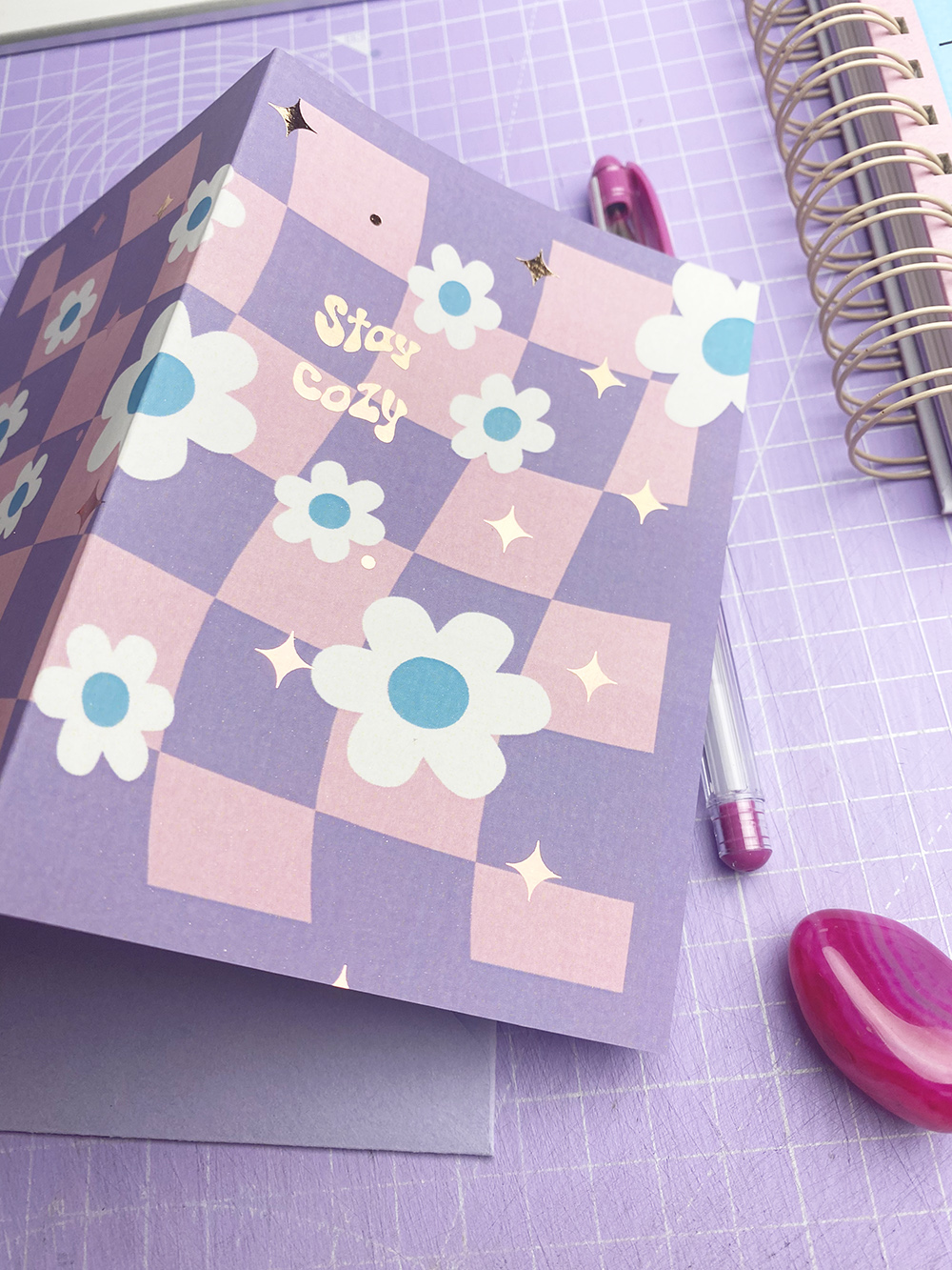 Cartão com padrão xadrez e flores, texto Stay cozy, sobre superfície lilás com caneta e caderno