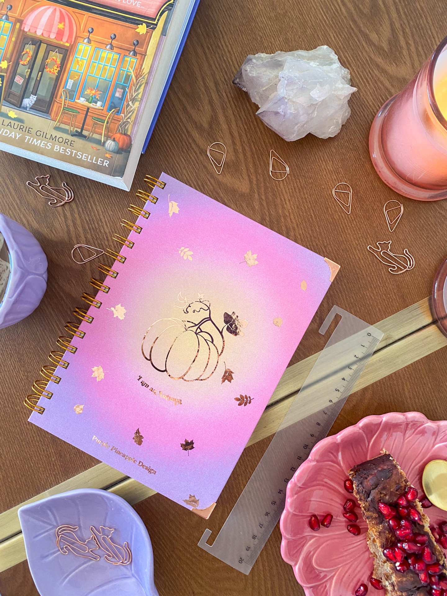 Caderno rosa lilás com abóbora dourada e texto, clips, cristal, régua, vela, pratos com sobremesa e livro sobre mesa de madeira