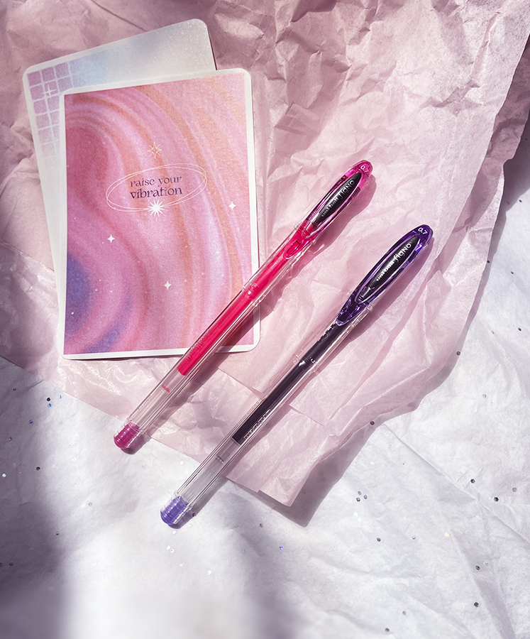 Dois cadernos pequenos coloridos e duas canetas gel rosa e roxa sobre papel de seda rosa.