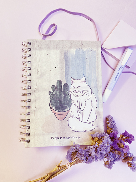 Caderno com capa ilustrada de cacto e gato, marcador e flores secas roxas