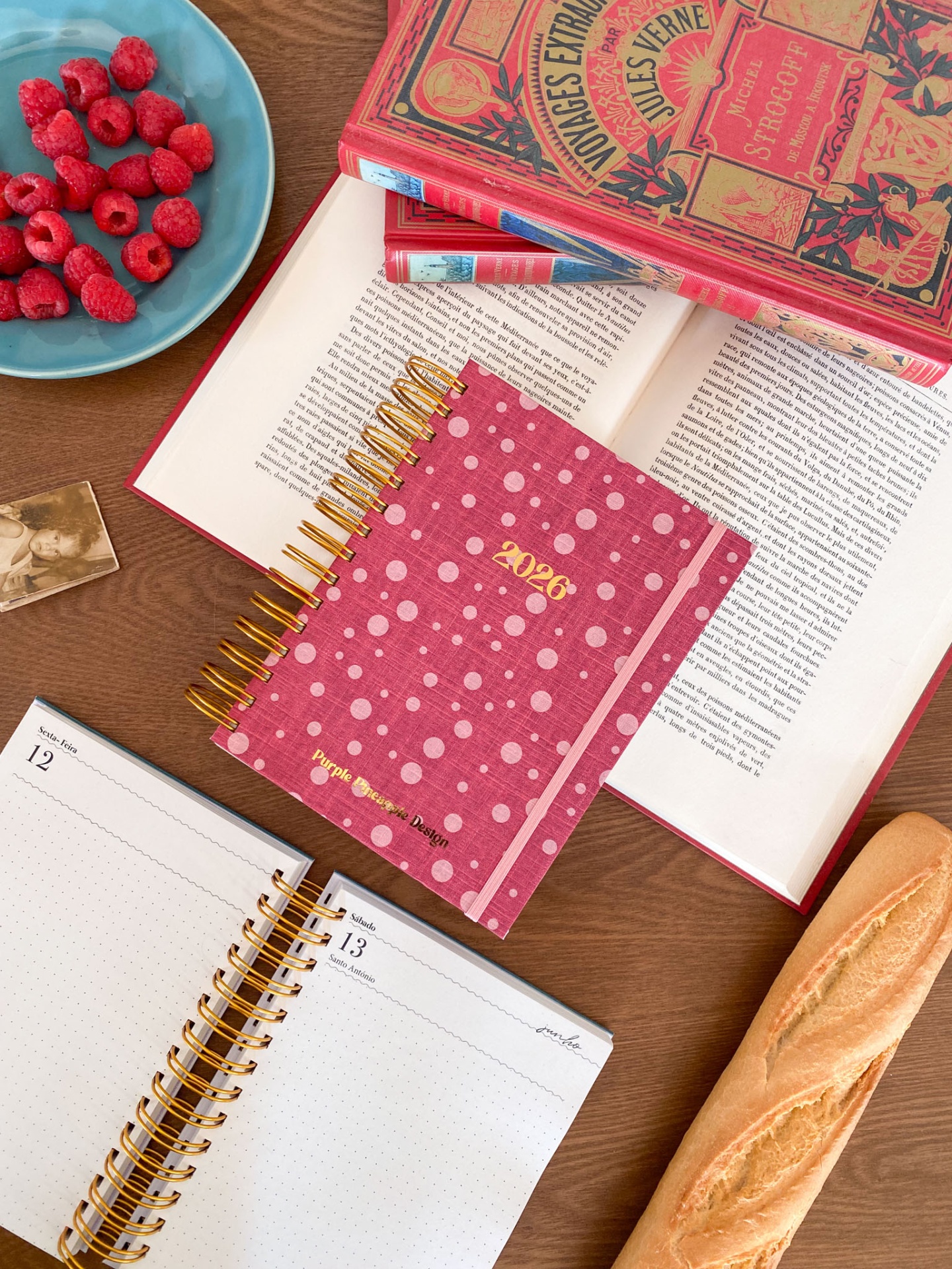 Agenda 2026 rosa com bolinhas, livros vermelhos, framboesas num prato azul e baguete numa mesa de madeira