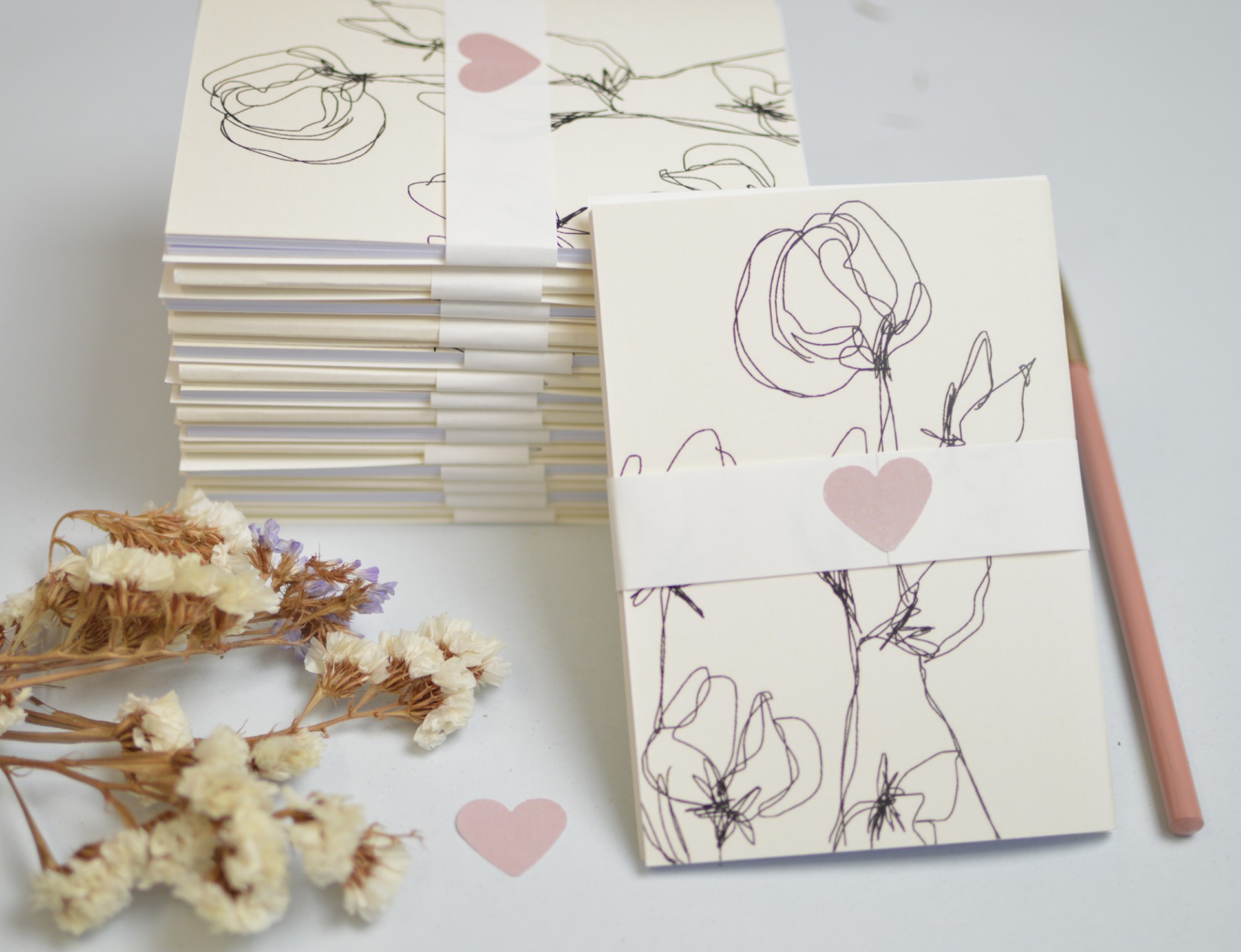 Cadernos com desenho floral e faixa com coração rosa ao lado de flores secas e lápis.