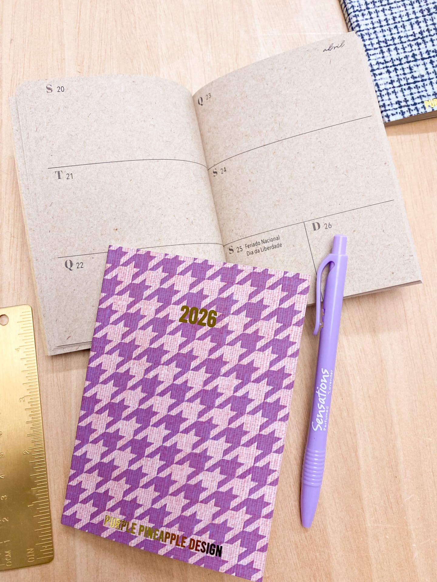 Agenda 2026 com padrão pied-de-poule roxo e rosa, caneta roxa e régua dourada sobre mesa clara
