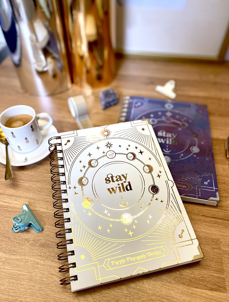 Dois cadernos espiralados decorativos com texto 'stay wild', chávena de café e clips sobre mesa de madeira