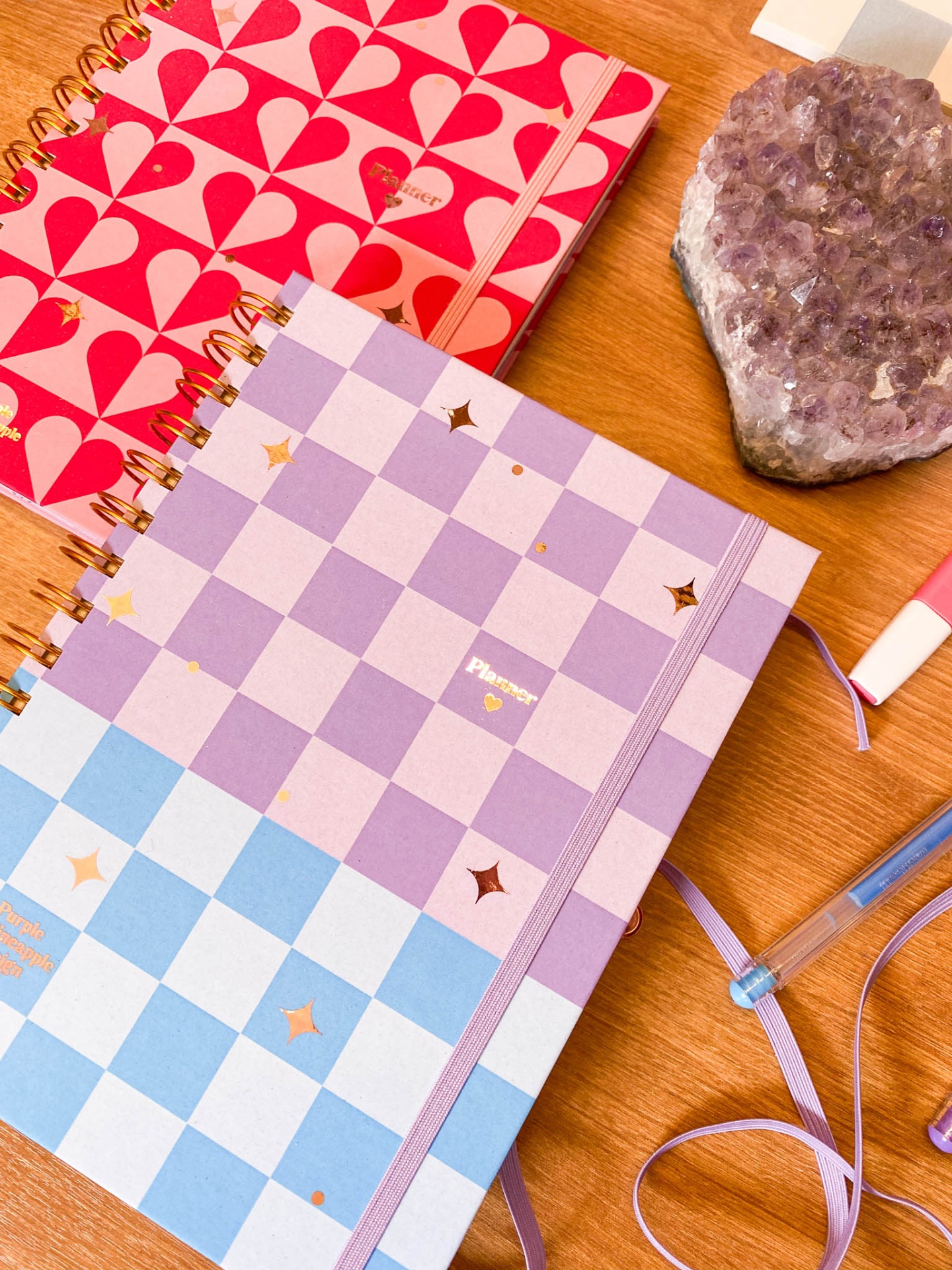 Dois cadernos coloridos espiralados com padrões distintos e elásticos, pedra ametista e canetas numa mesa de madeira