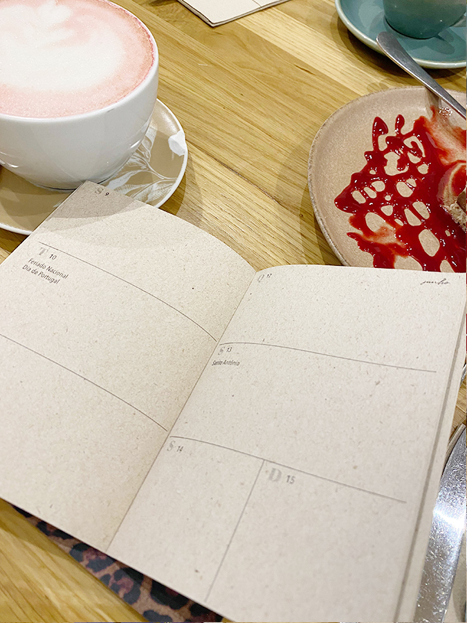 Livro aberto com datas, chávena branca com bebida rosa, pires castanho com molho vermelho, mesa de madeira