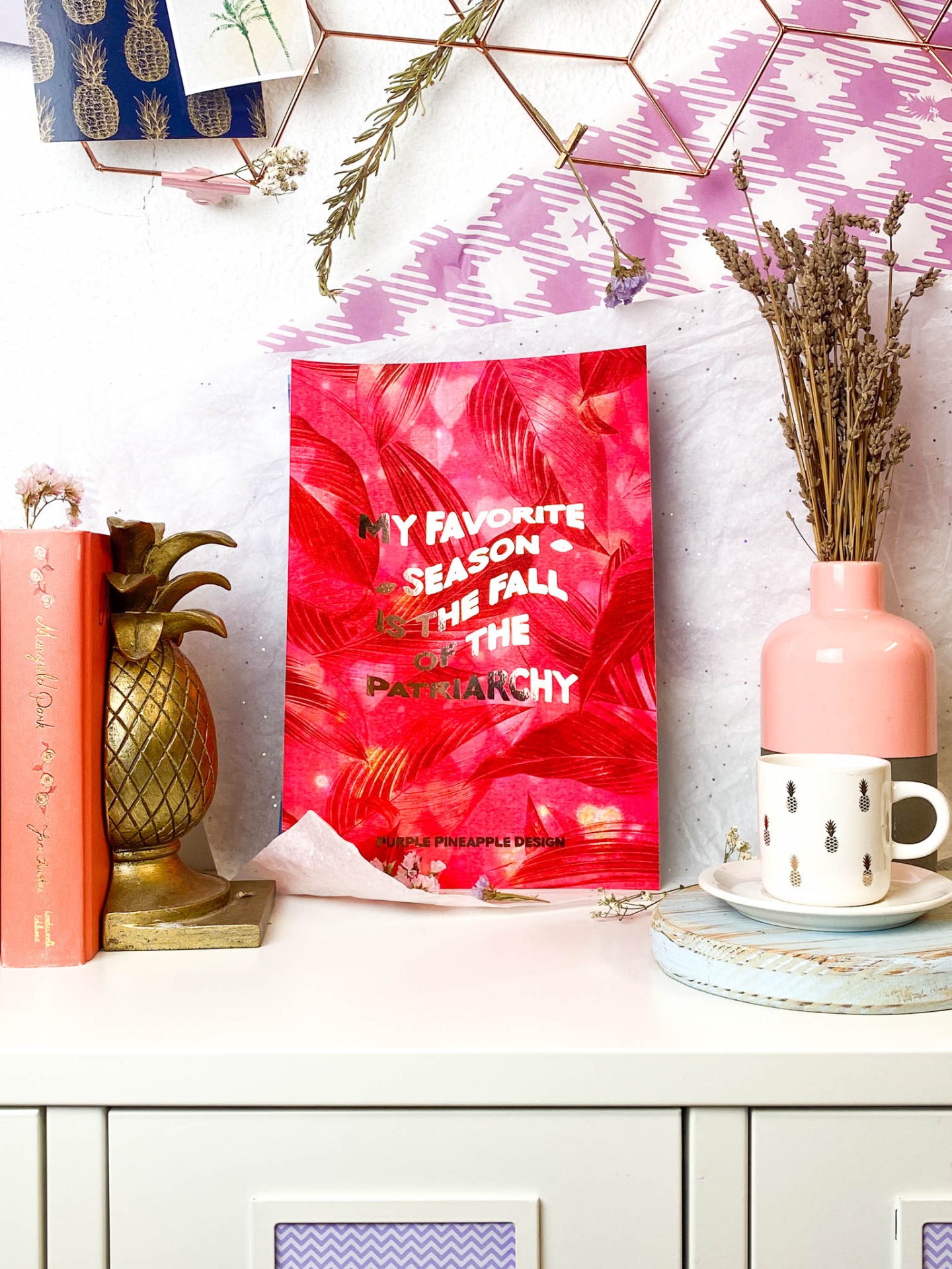 Decoração com livro laranja, suporte abacaxi dourado, saco vermelho com texto, jarra rosa com flores secas, chávena branca e tecido xadrez rosa e branco