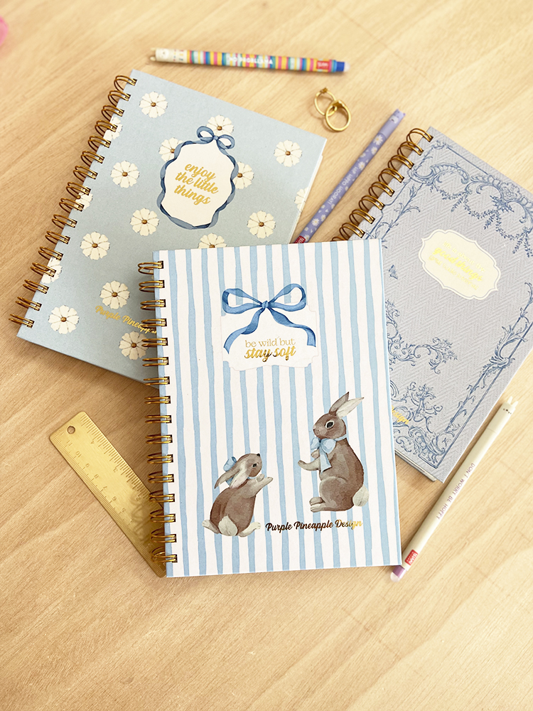 Três cadernos espiralados com capas azuis decoradas e texto em inglês sobre uma mesa de madeira