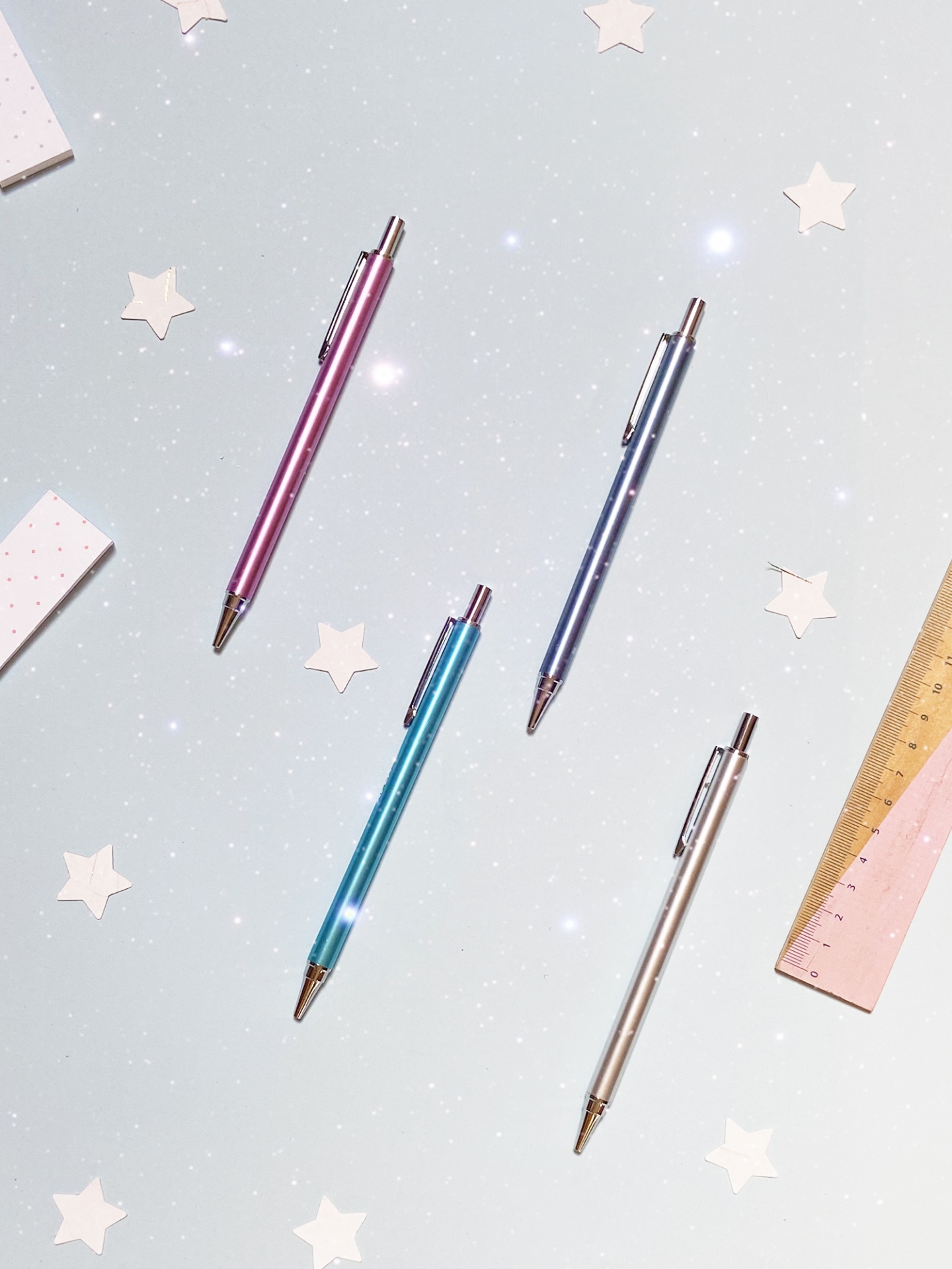 Quatro canetas metálicas coloridas sobre superfície branca com estrelas e réguas ao lado