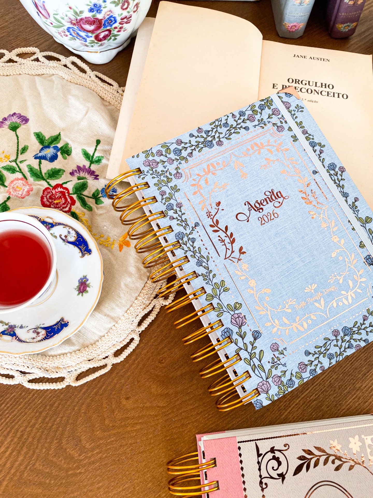 Caderno floral azul com texto Agenda 2026 na mesa com chávena de chá vermelho e livro aberto