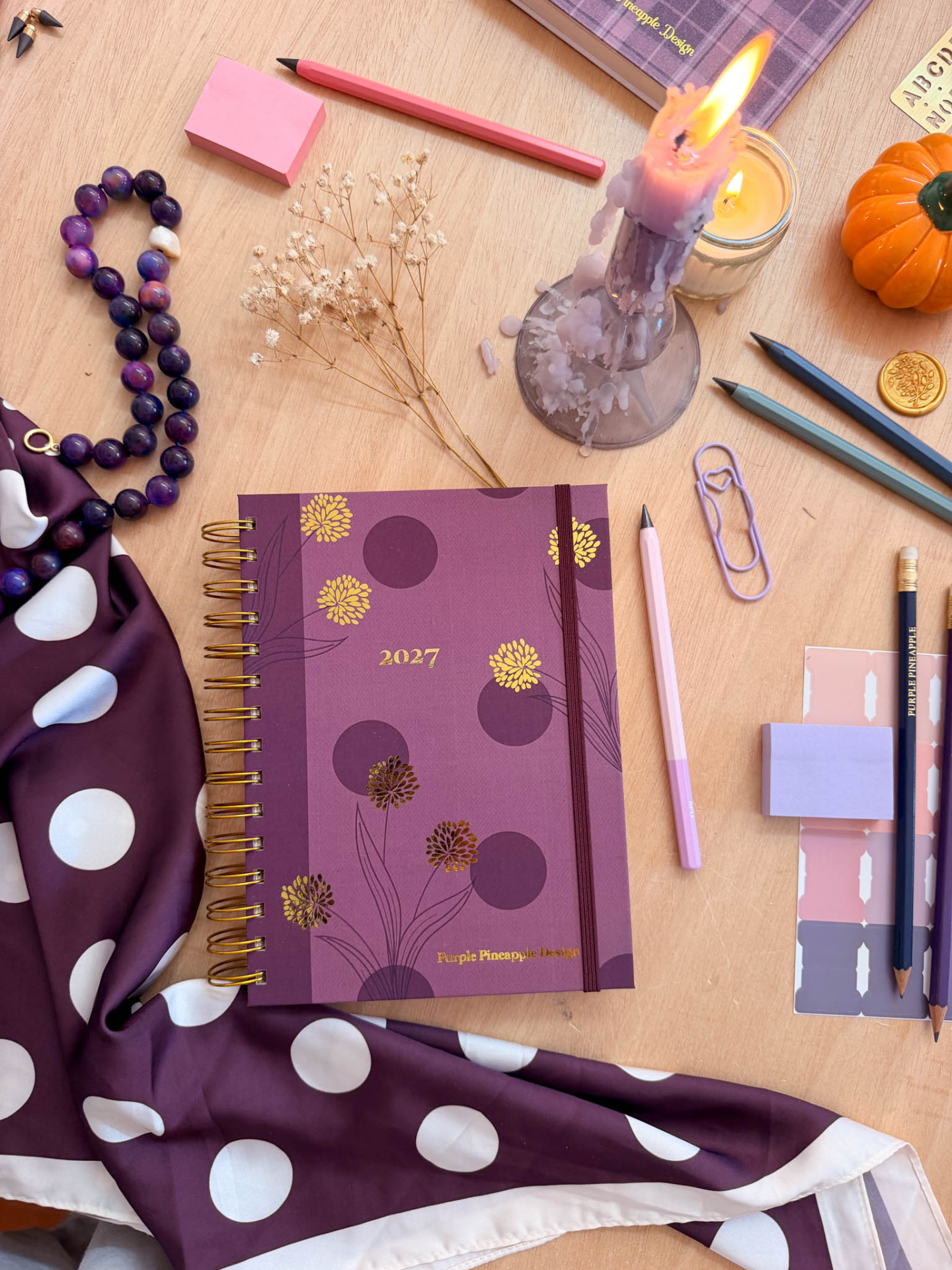 Agenda 2027 roxa com flores douradas, canetas coloridas, vela acesa, lenço roxo com bolinhas, e outros artigos de papelaria em mesa de madeira.