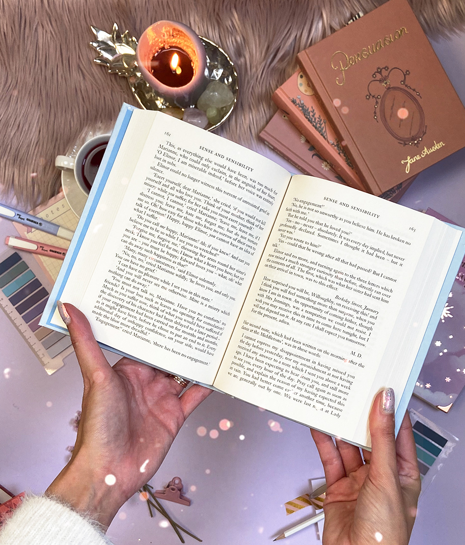 Livro aberto a texto em inglês, mãos a segurá-lo, livros cor de rosa, vela acesa e chávena de chá num ambiente acolhedor