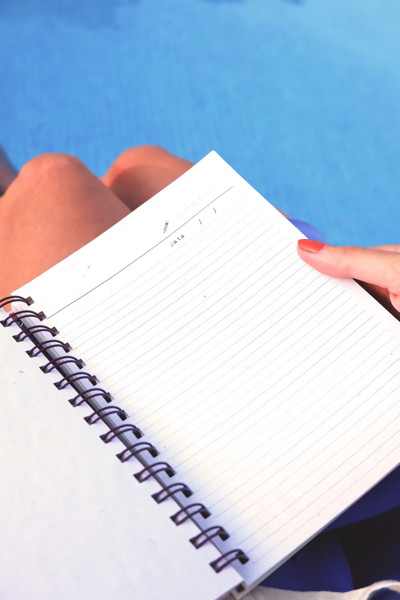 Caderno espiral branco com linhas e campo para data, mão a segurar, pernas e piscina ao fundo