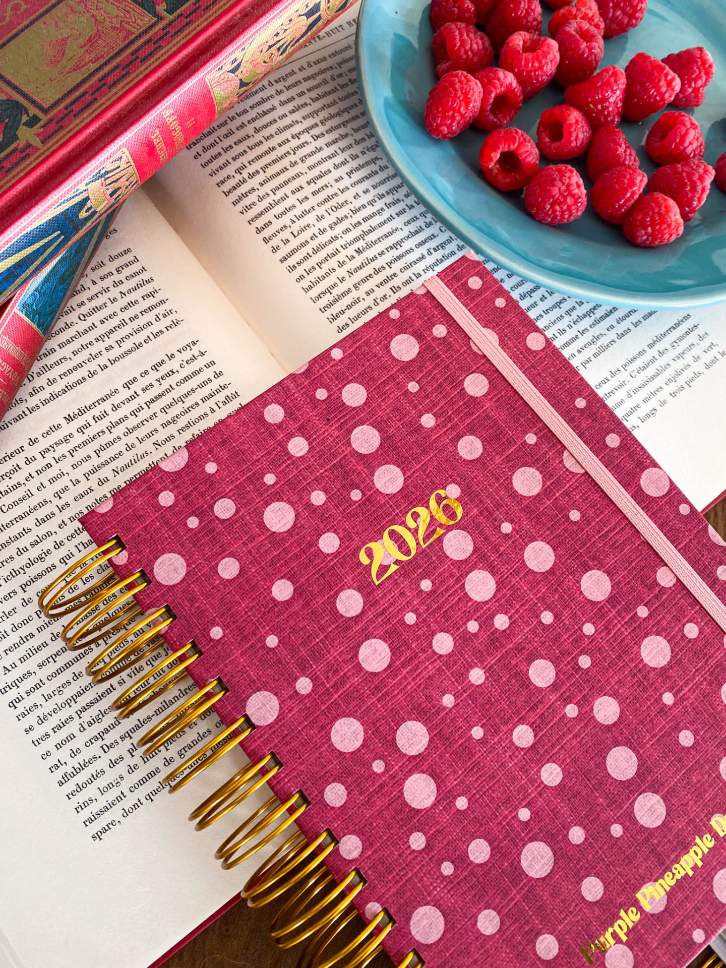 Agenda rosa com bolas brancas e texto dourado sobre livros e prato azul com framboesas vermelhas