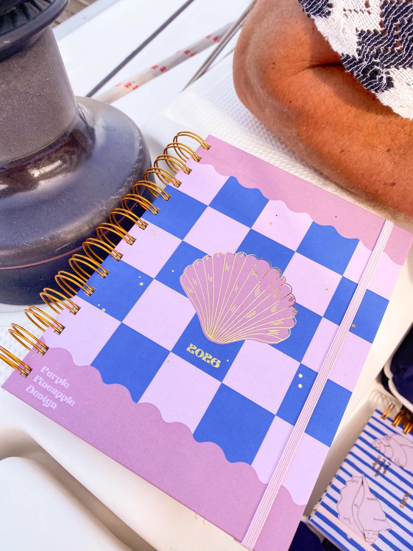 Caderno rosa e azul com concha dourada e elástico rosa numa mesa ao lado de um vaso e tecido
