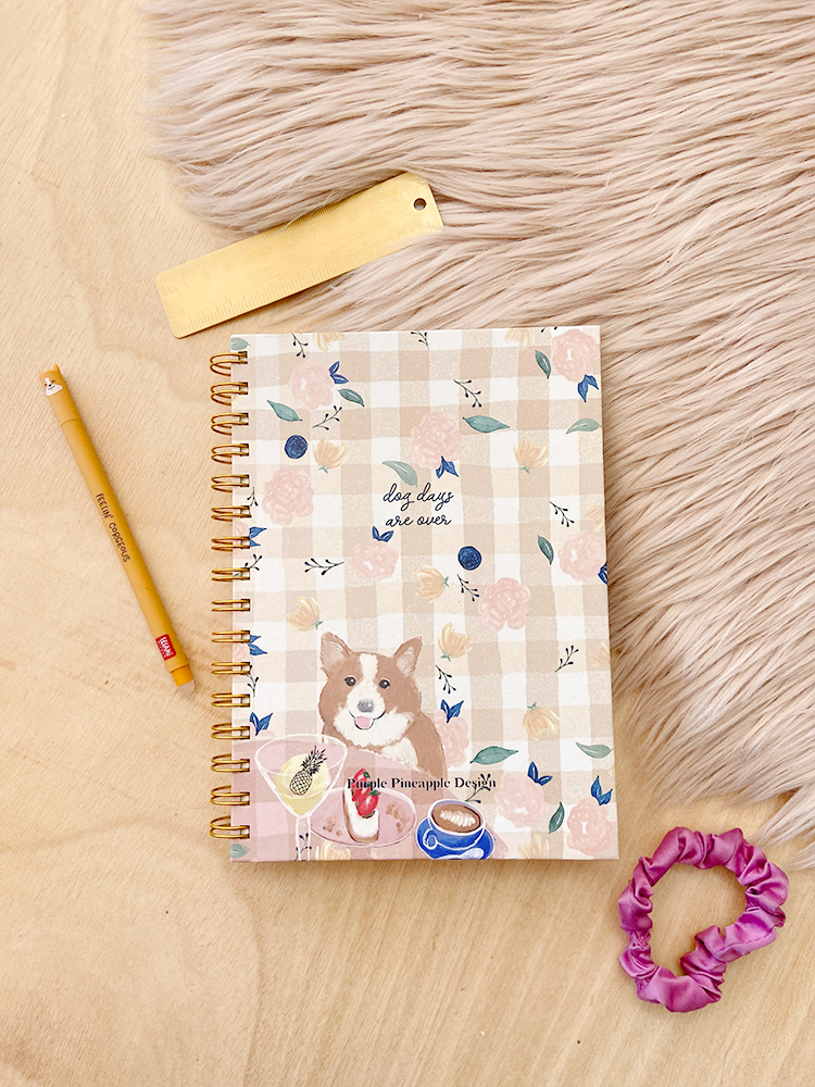 Caderno espiral com capa estampada e itens de papelaria dispostos sobre mesa de madeira.