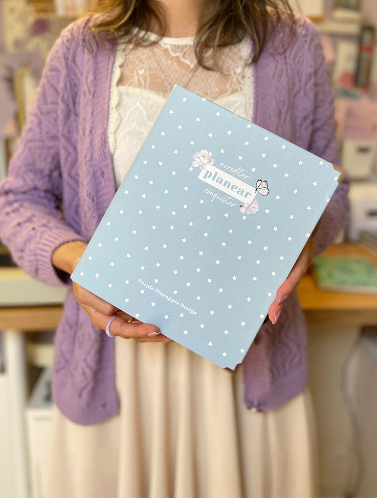 Pessoa a segurar caixa azul clara com estrelas brancas e texto, vestindo casaco roxo e blusa branca