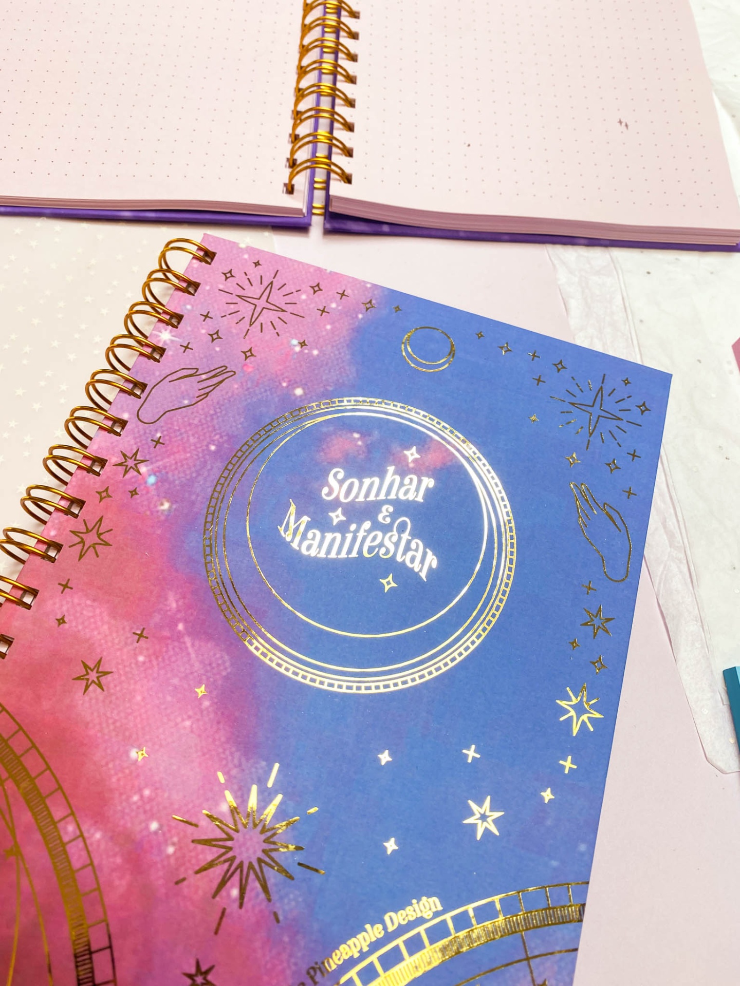 Cadernos com capa dura e espiral dourada, capa azul e rosa com texto e símbolos dourados