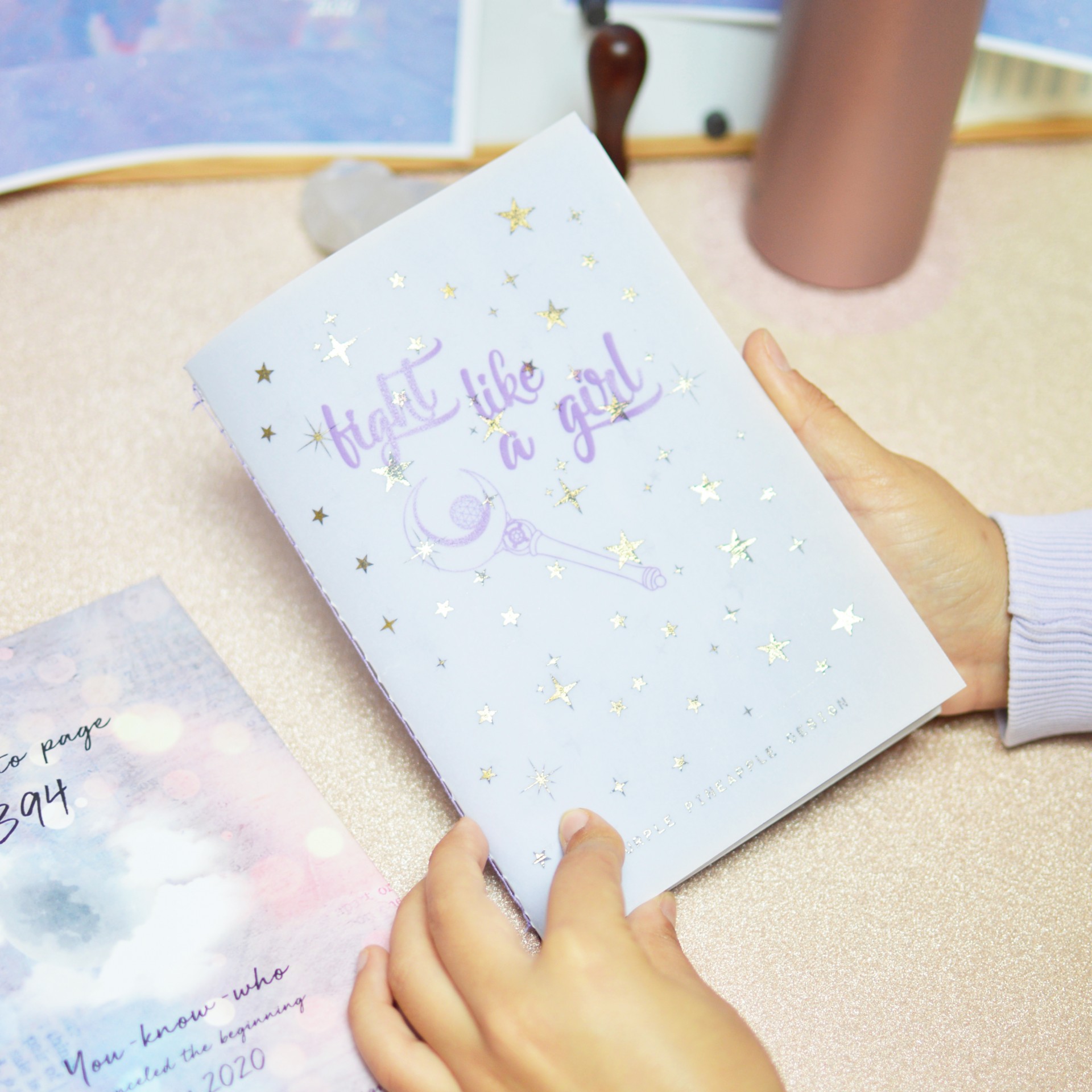 Caderno branco com estrelas douradas e texto lilás segurado por mãos sobre mesa clara