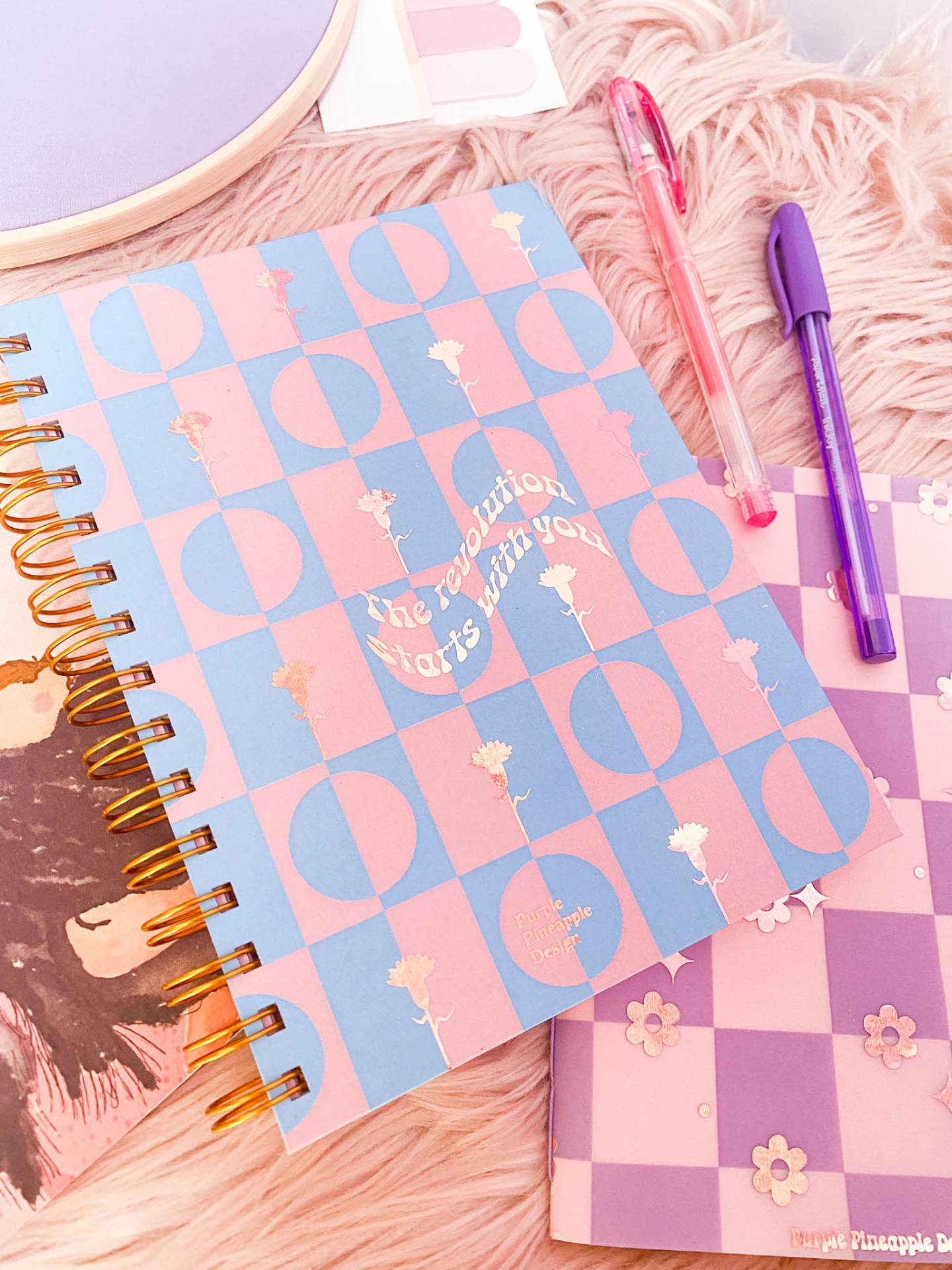 Cadernos coloridos com padrões quadriculados e flores, duas canetas, em superfície felpuda rosa.