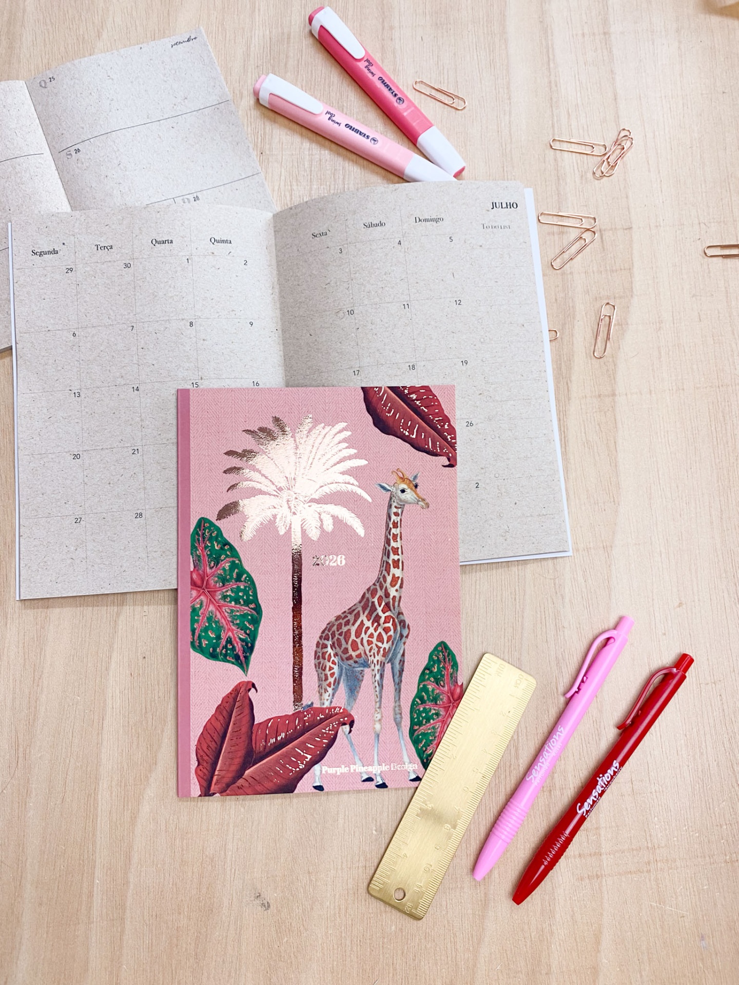 Caderno rosa com girafa e plantas, calendário aberto, canetas e marcadores rosa e vermelho, régua dourada e clips dourados