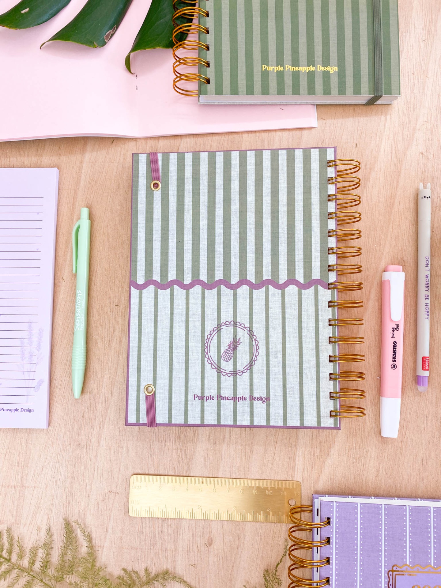Cadernos e material de escritório organizados sobre mesa de madeira com caderno verde riscado com texto Purple Pineapple Design