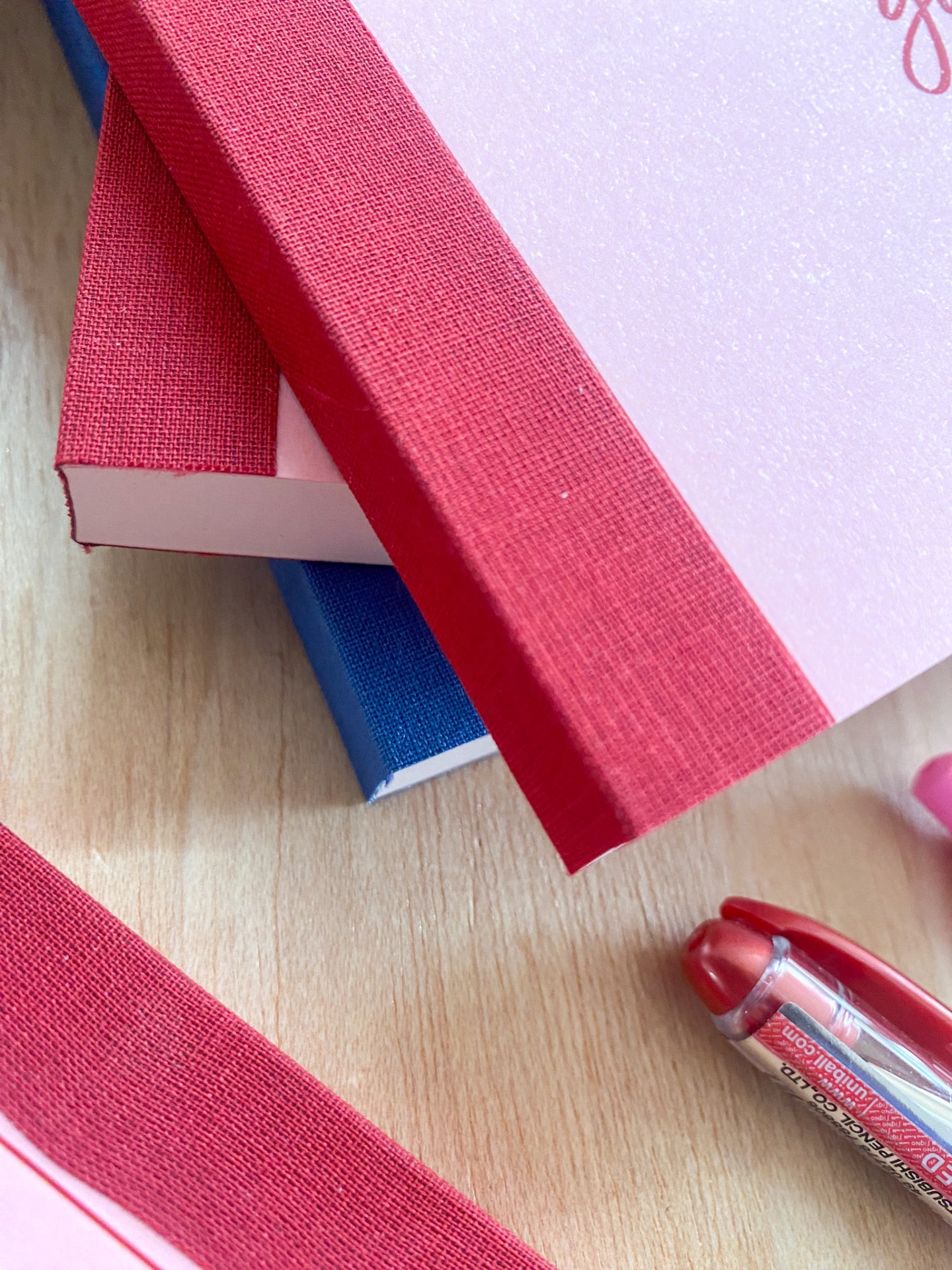 Três cadernos com lombadas vermelhas e capas rosa e azul, caneta vermelha num fundo de madeira clara