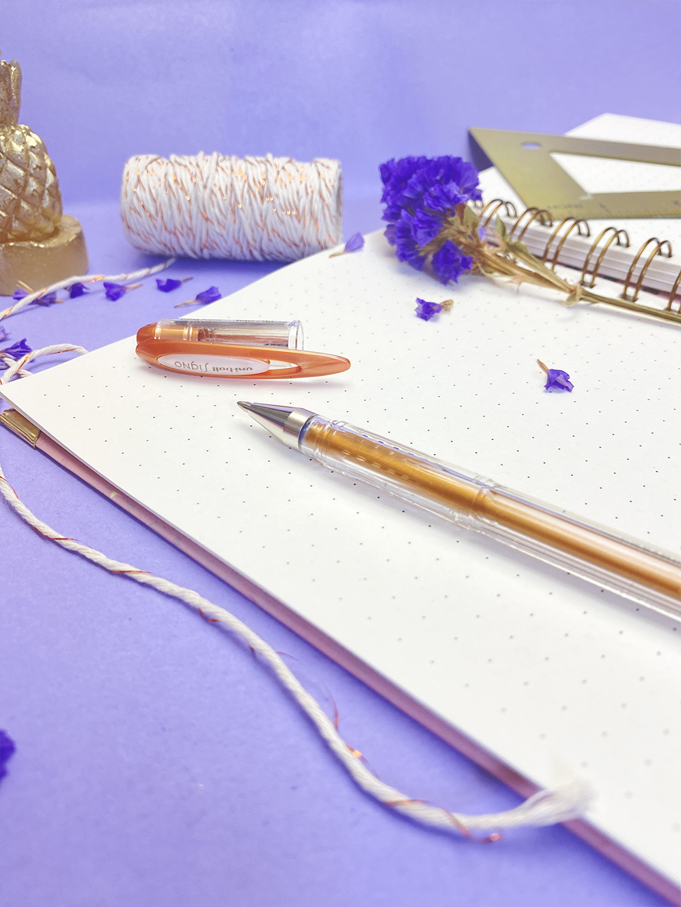 Bloco de notas espiralado com caneta laranja, cordão branco dourado e flores roxas em fundo lilás