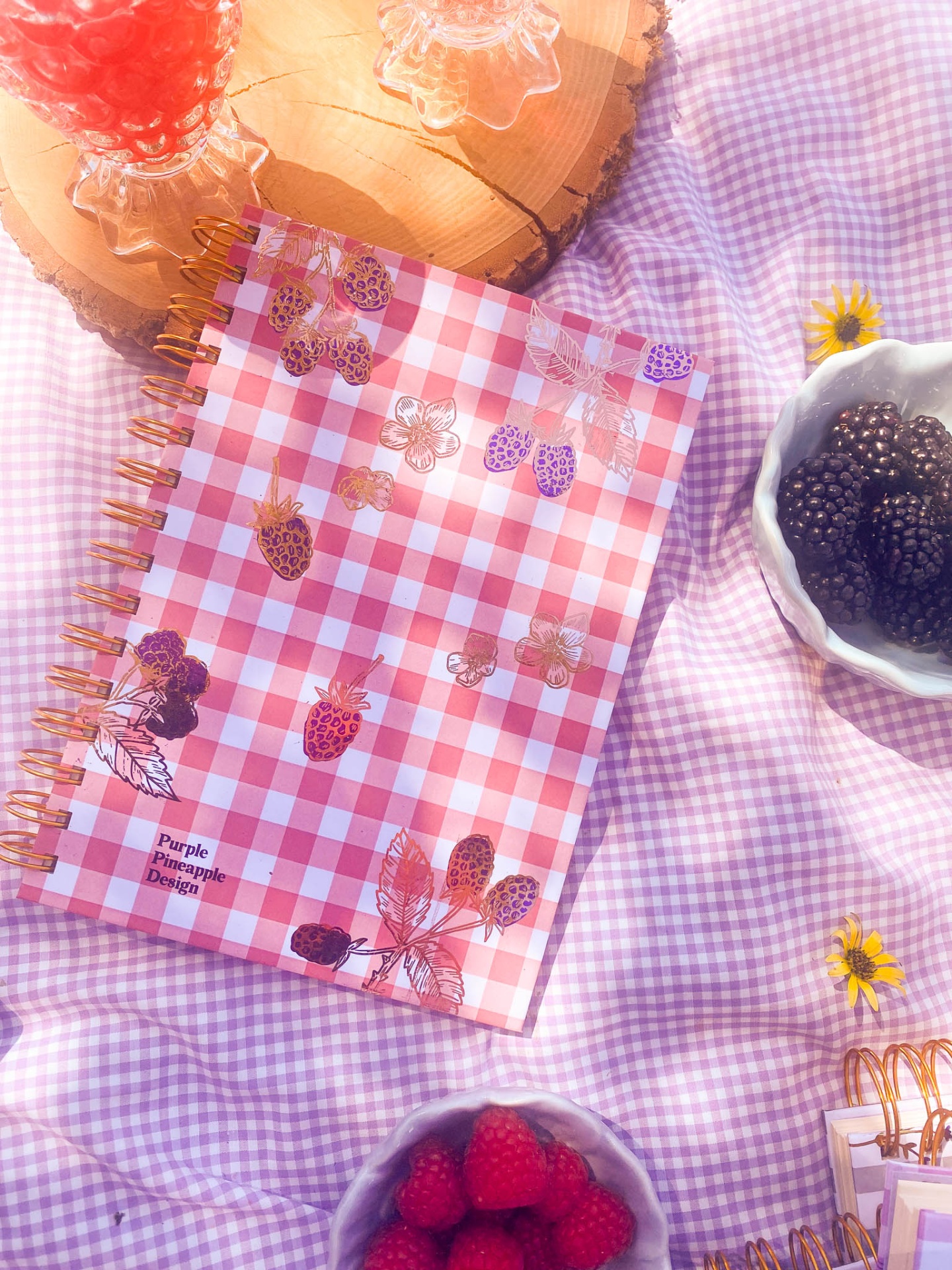 Caderno xadrez com frutas e texto Purple Pineapple Design sobre tecido lilás xadrez com tigelas de frutas e copos