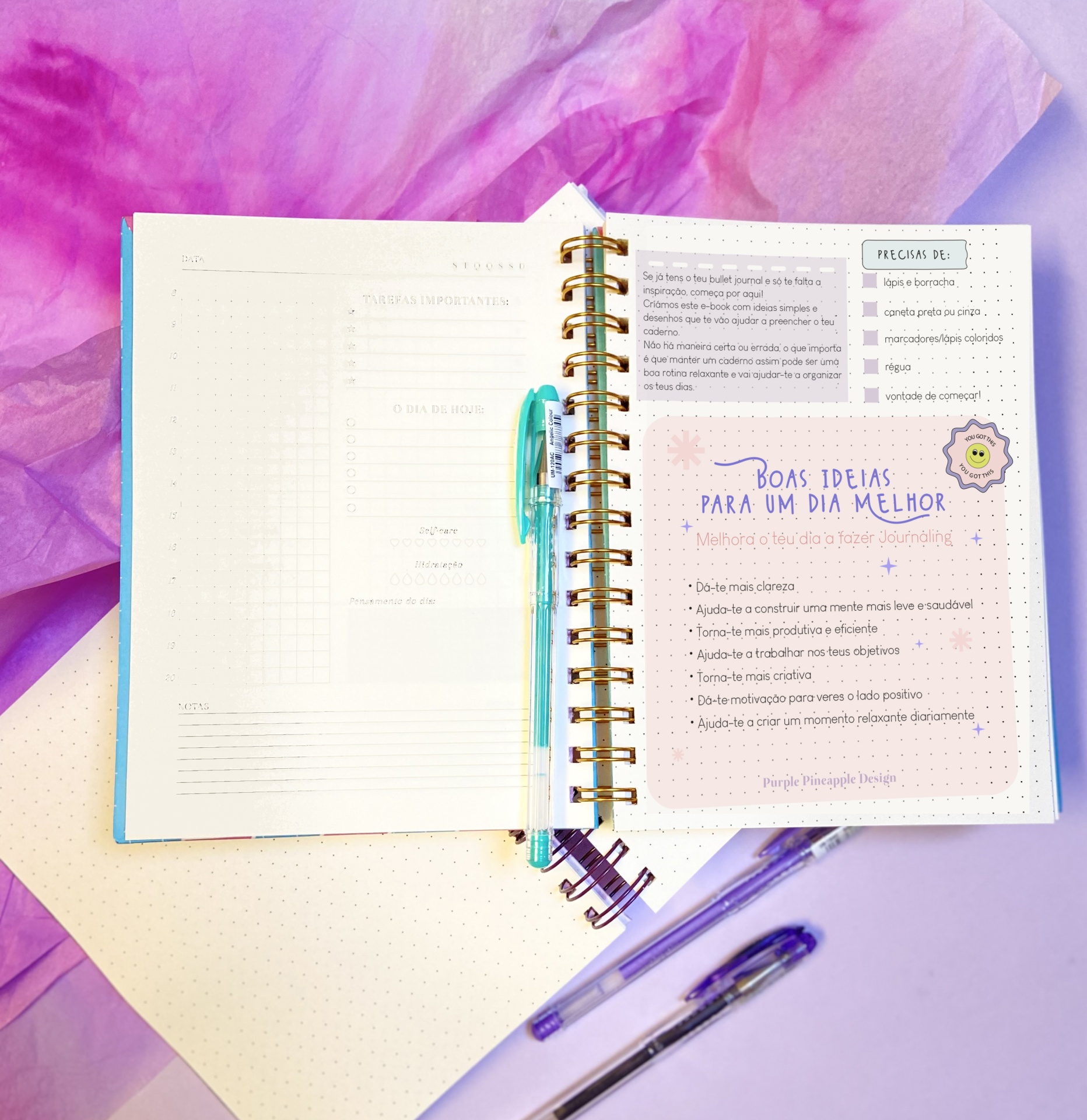 Agenda aberta com texto em português e caneta azul em cima, sobre fundo lilás e tecido rosa