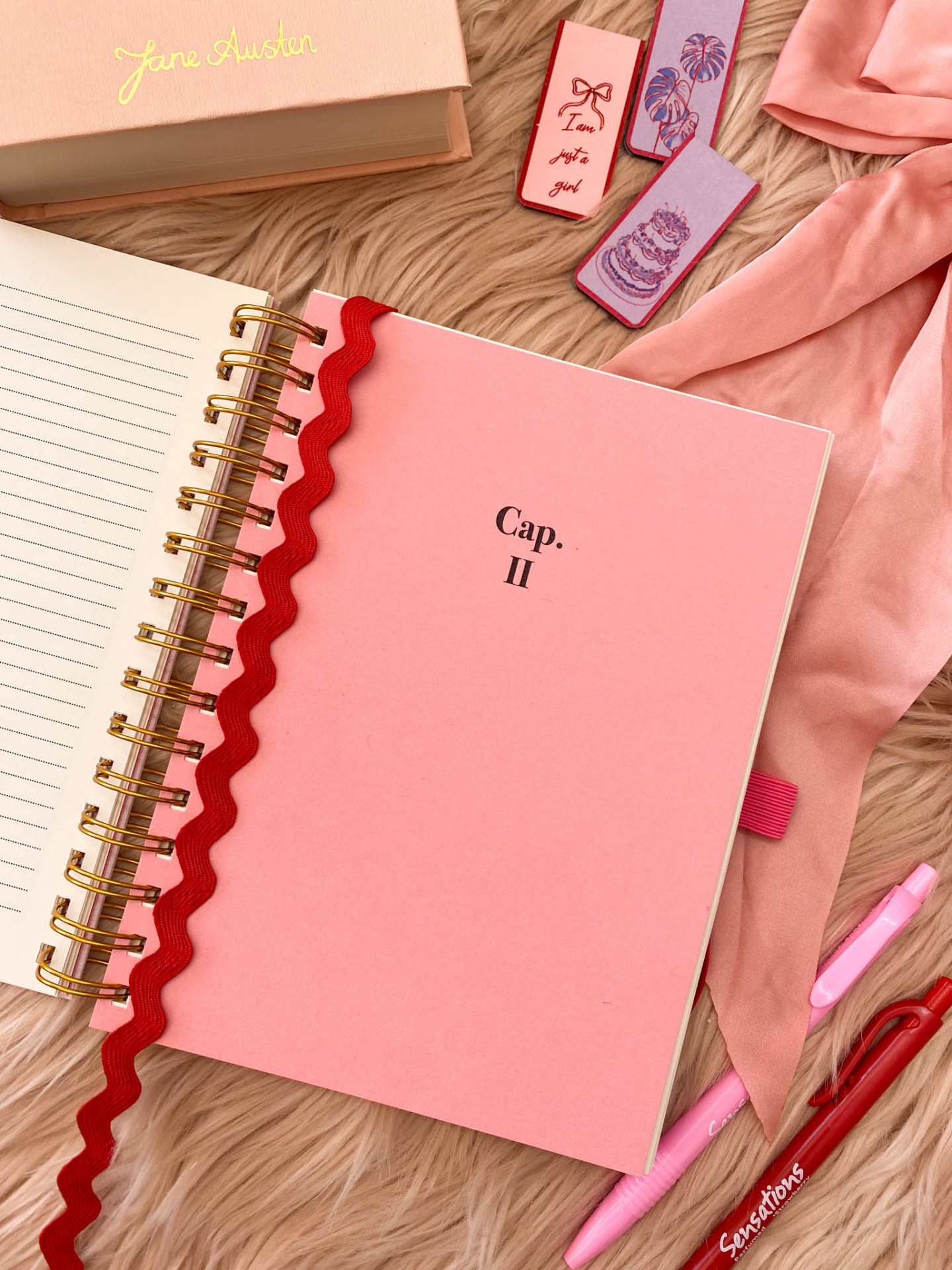 Caderno rosa com capa com texto 'Cap. II' e acessórios de escrita sobre superfície peluda