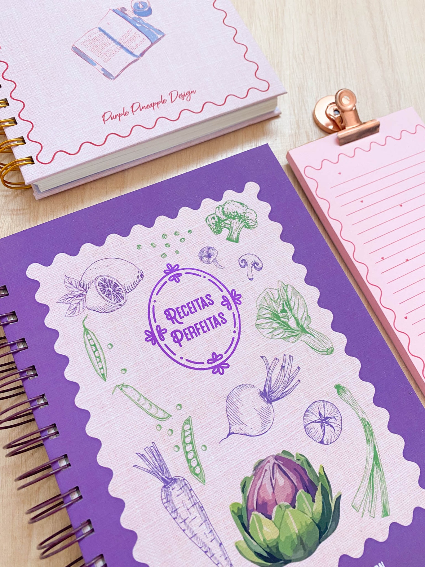 Três cadernos coloridos, um roxo com vegetais desenhados e texto, outro rosa claro com espiral dourada, e um bloco rosa com clipe metálico.