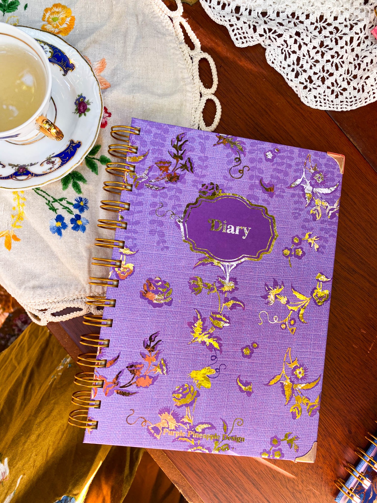 Caderno diário roxo com flores douradas sobre mesa de madeira com toalha bordada e chávena de chá