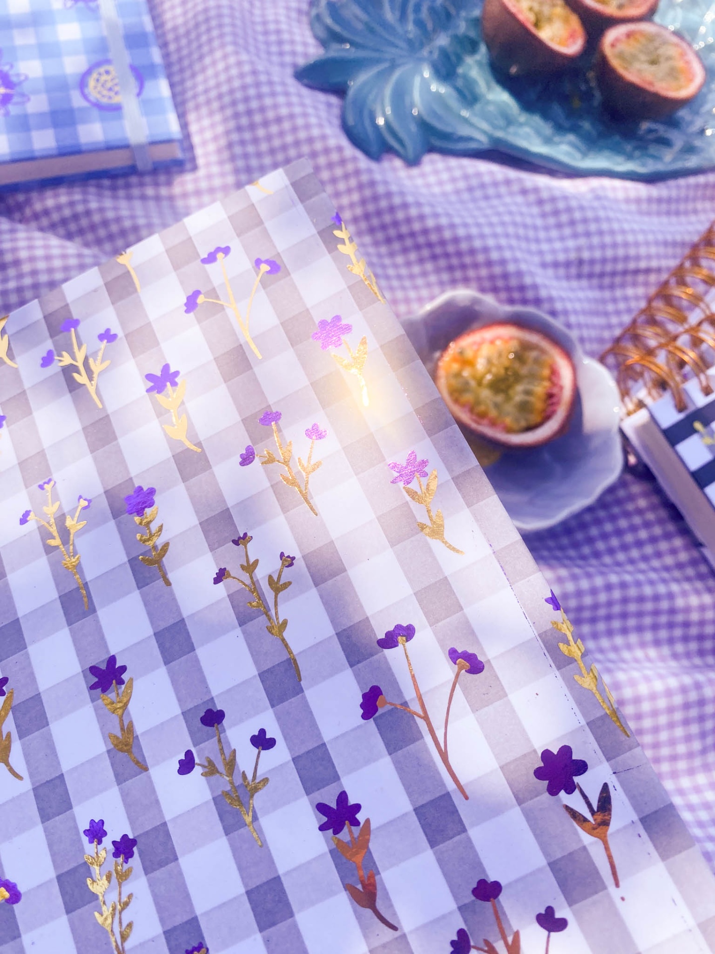caixa decorativa com padrão xadrez branco e lilás e flores douradas e roxas sobre mesa lilás com maracujás e prato azul