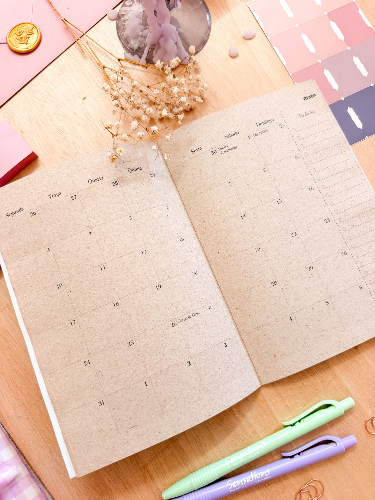 Agenda mensal em papel reciclado com decoração de flores secas, canetas Sensations verde e lilás, carimbo dourado e paleta de cores rosa e lilás, sobre mesa de madeira.