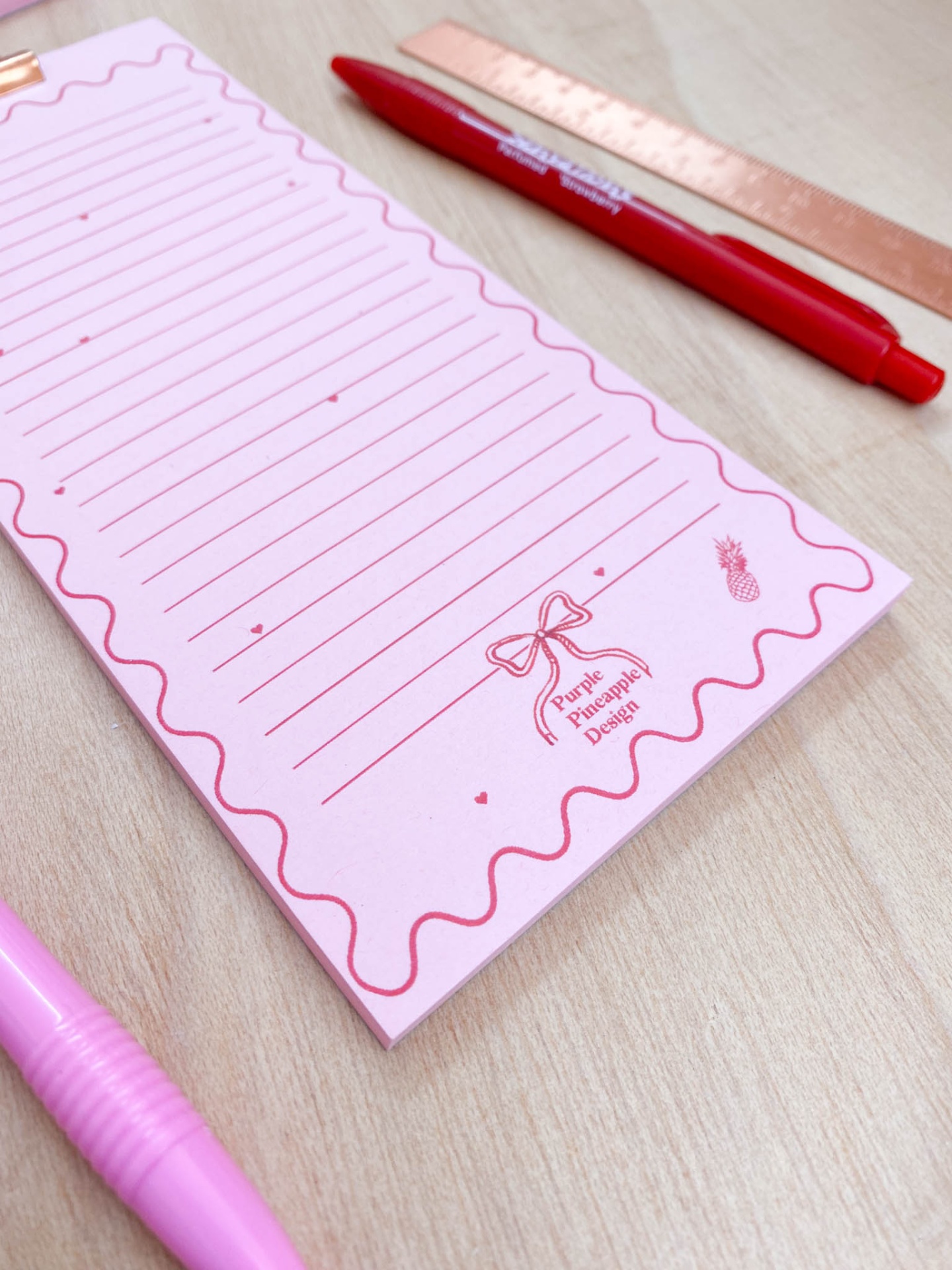 Bloco de notas rosa com canetas e régua sobre mesa de madeira.