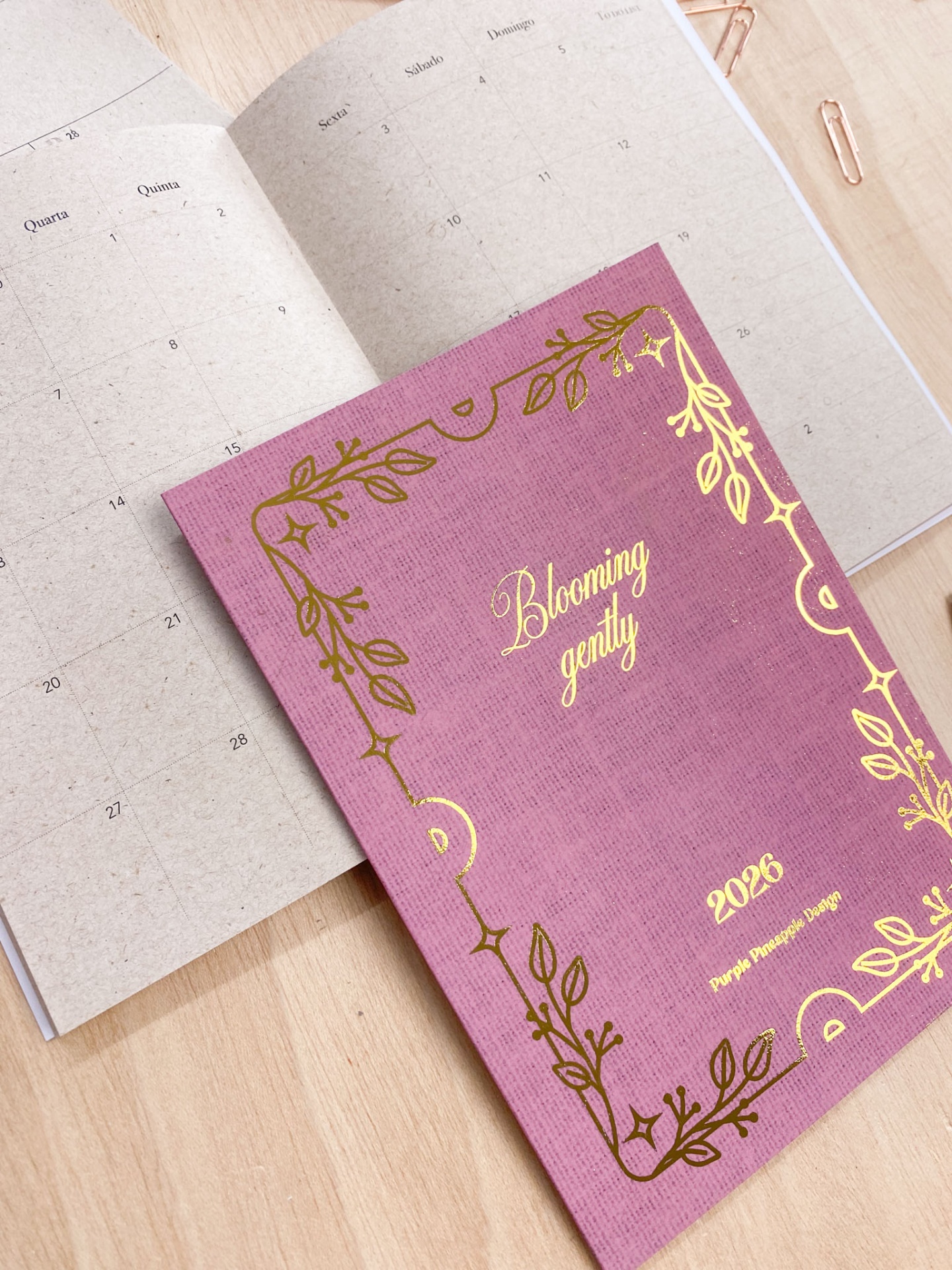 Planner rosa 2026 com detalhes dourados aberto em página de calendário monthly.