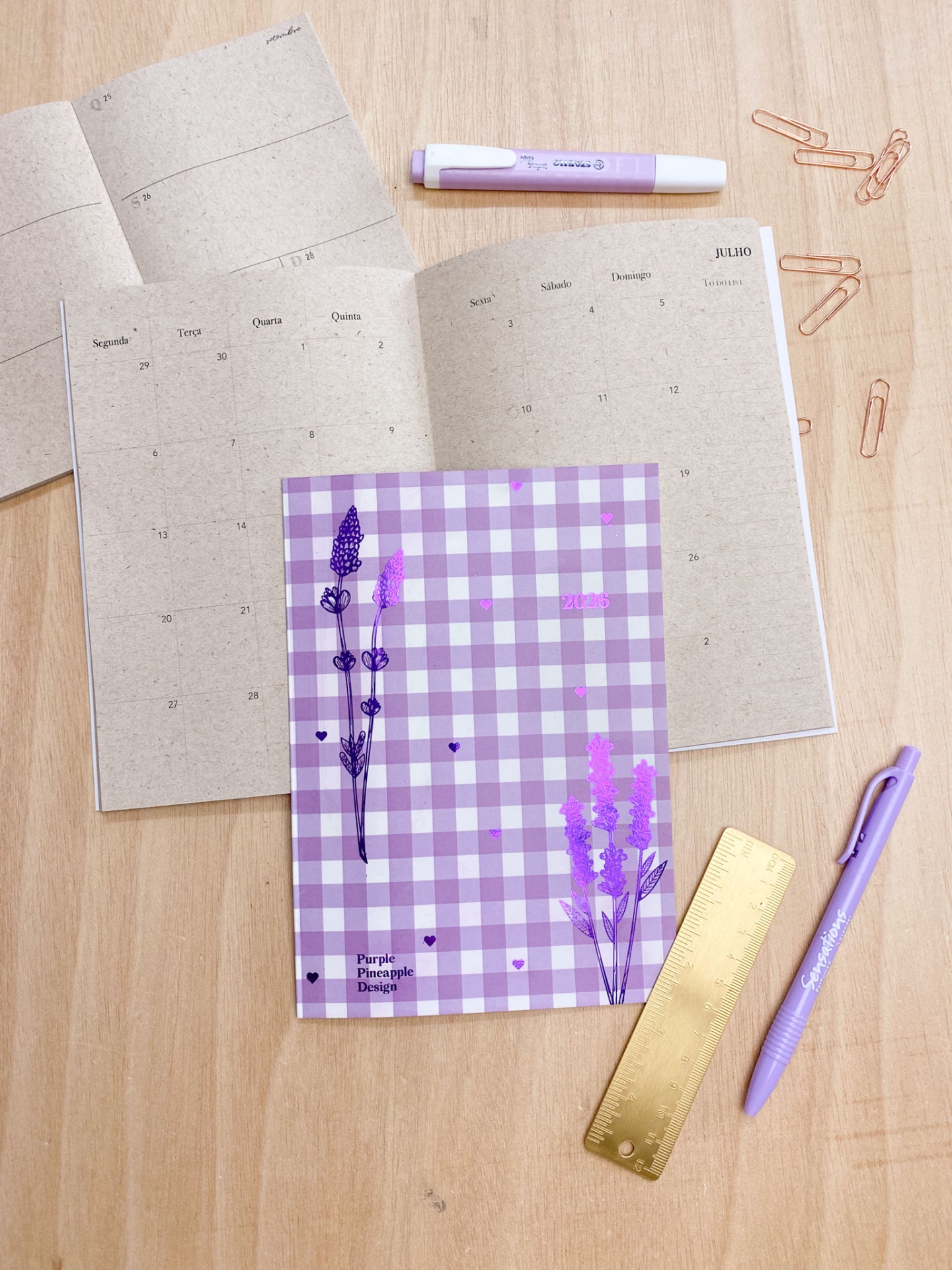 Planner aberto com capa xadrez lilás e objetos de escritório lilás e dourado sobre mesa clara